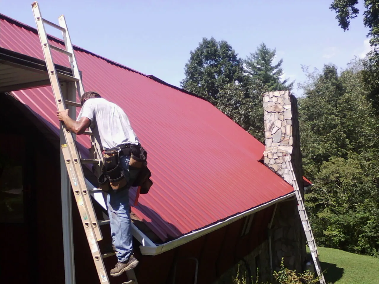 Roofer on a ladder beside a red metal roof and stone chimney — Soffit & Fascia Repair by Roofing Brothers in Falmouth